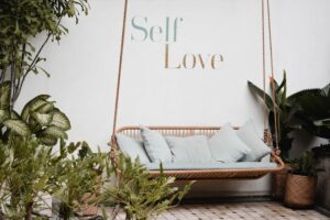 outdoor Sofa brow wicker swing with soft white cushions, surrounded by plant - with text "Self Love" written on the wall.
