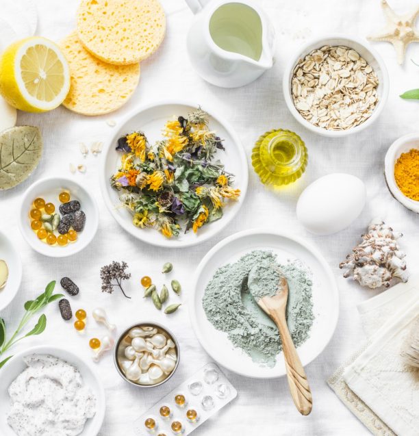 Home beauty products - clay, oatmeal, coconut oil, turmeric, lemon, scrub, dry flowers and herbs, sponges, soap, facial brush on light background, top view. Skin youthfulness, beauty concept. Flat lay